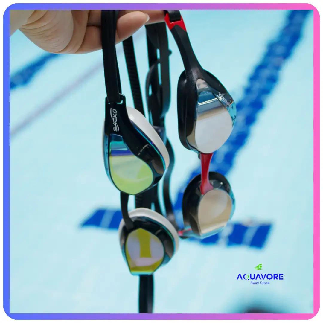 Carina Mirror swimming goggles with interchangeable lenses hanging by straps, showcasing reflective and tinted lens options. Aquavore logo visible in corner, pool lanes in background.